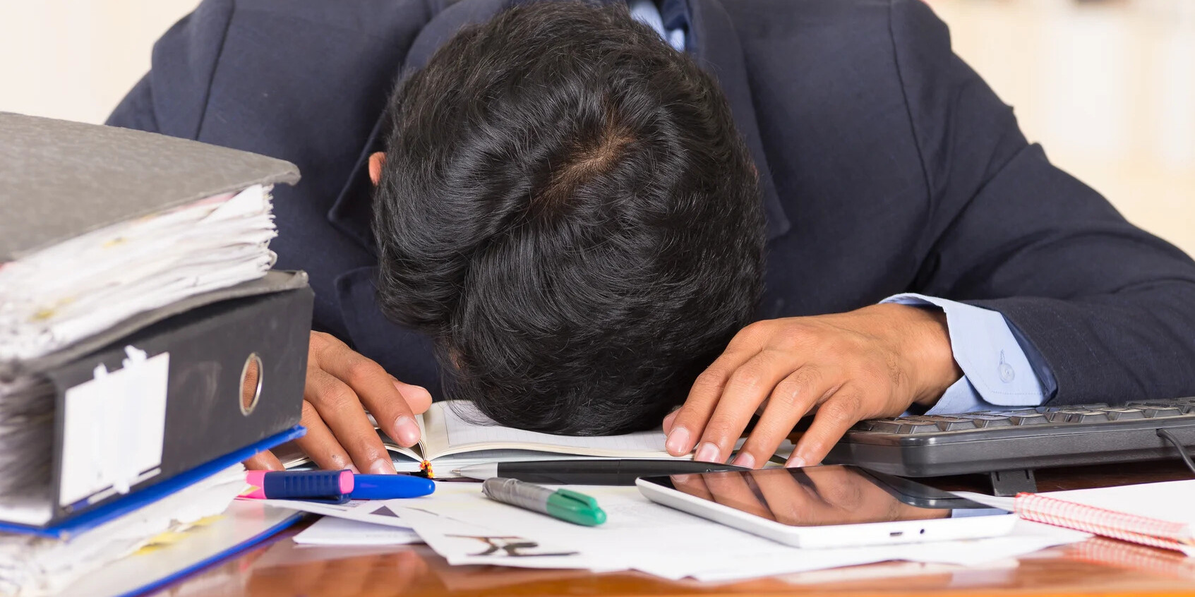 man with head on desk with paperwork and binders